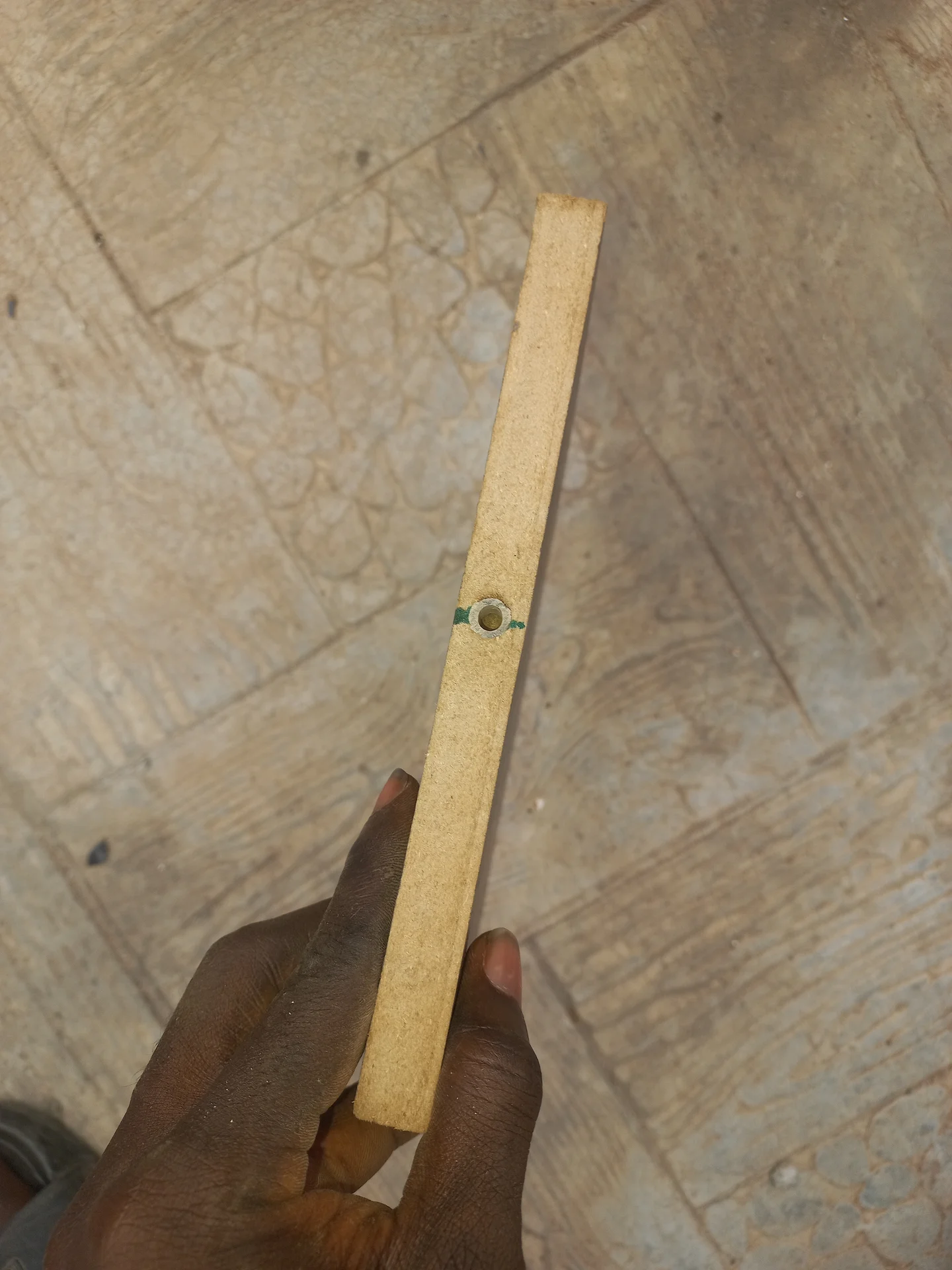 A narrow wooden strip held in a hand showing a small hole drilled through it, used as a bracket or mounting piece for a sensor