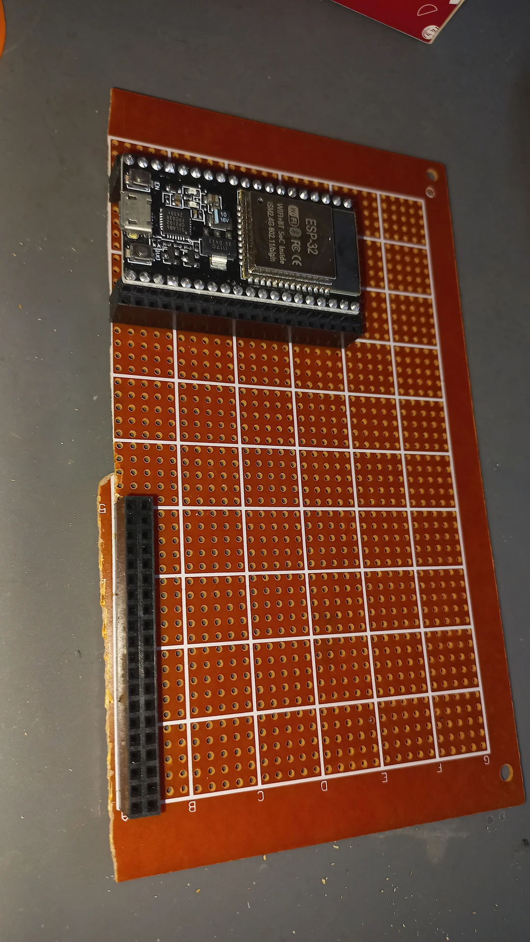 Wider view of the perfboard with the ESP32 positioned at the top and a pin header strip along the left edge, before any wiring