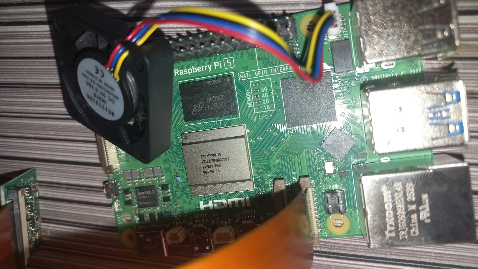 Angled top view of the Pi 5 board showing the blower fan, chip area, GPIO header and the Raspberry Pi 5 label, with the camera ribbon visible at the edge