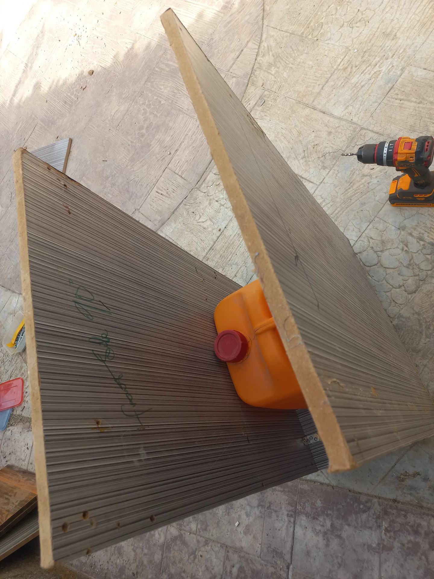 Two corrugated side panels propped together on the ground during construction, with a jerry can used as a weight and a power drill nearby