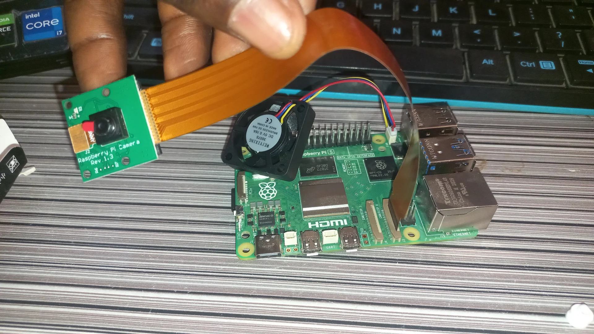 Pi Camera Module held up in a hand connected via ribbon cable to the Pi 5 below, showing the lens and the assembled system together