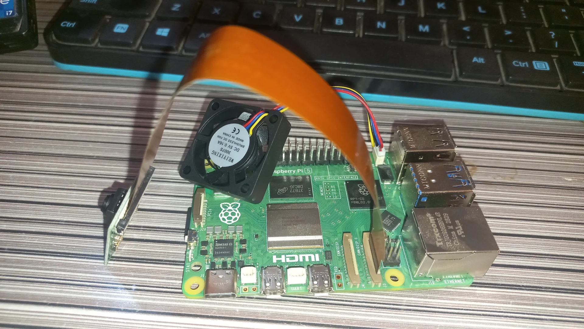 Angled front view of the Pi 5 with the blower fan and the camera ribbon cable arching over the board, showing the HDMI port and GPIO header