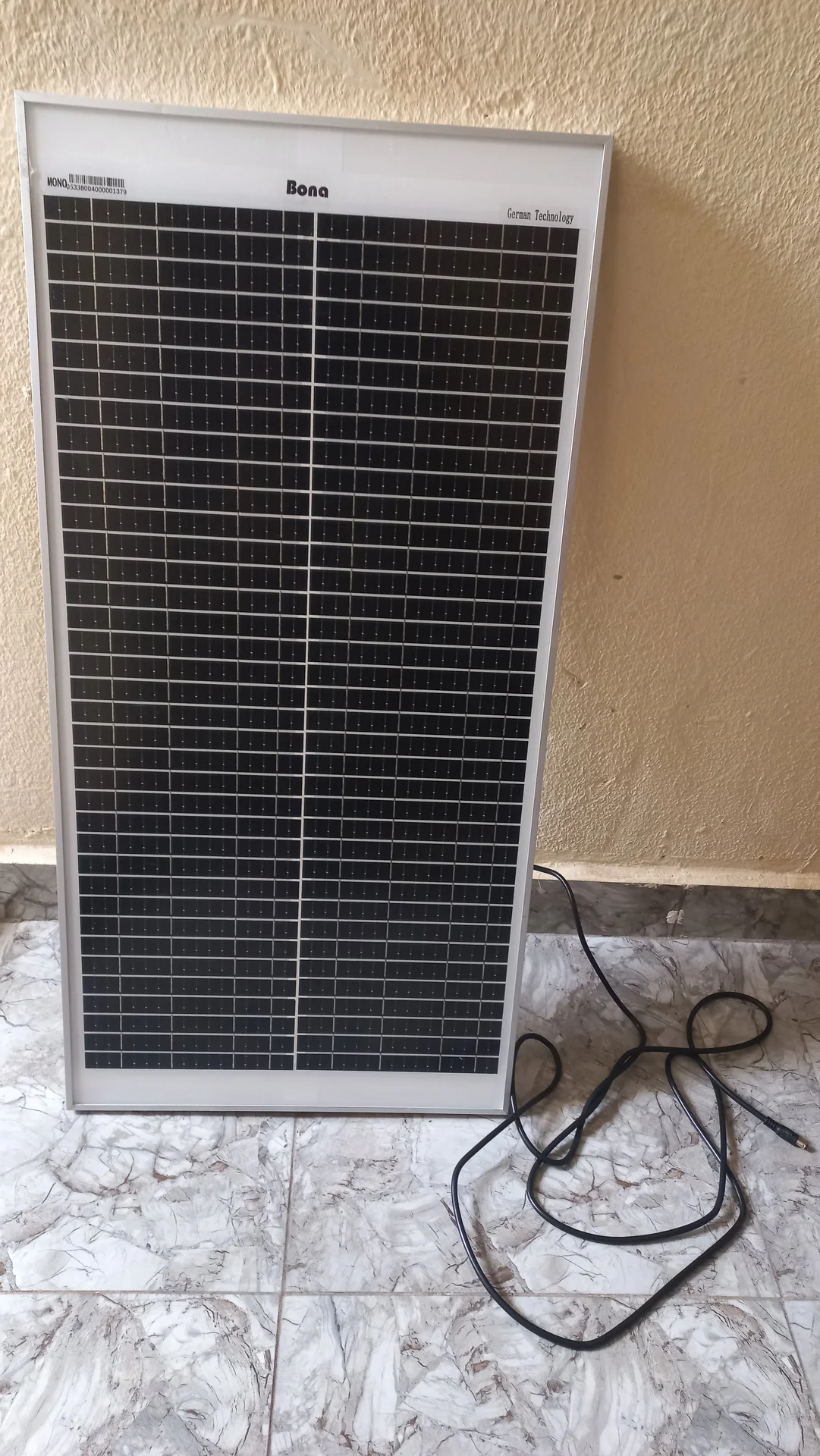 50 W Bona solar panel standing upright against a wall, front face showing the cell grid and aluminium frame, with cable on the floor